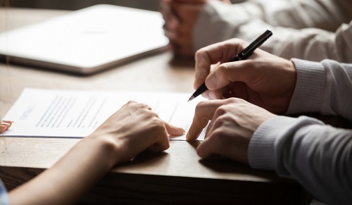 A document on a table, one person pointing to it as another signs it with a pen