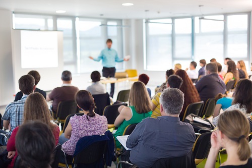 A room full of people sat at desks, with one person presenting at the front